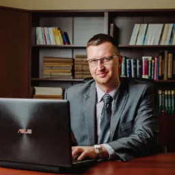 Mężczyzna w garniturze i okularach siedzi za biurkiem z laptopem Asus, w tle regał z książkami.