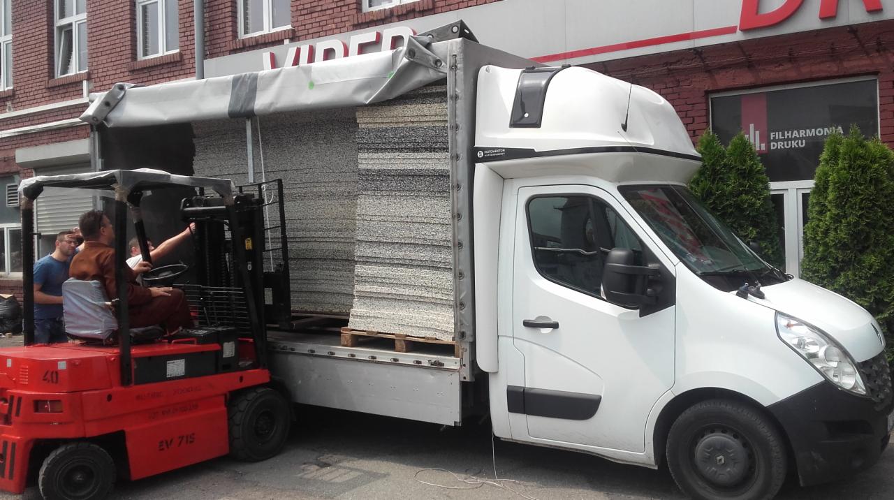 Czerwony wózek widłowy podnosi paletę z warstwami szarej pianki do białego samochodu dostawczego z otwartą plandeką. W tle budynek z czerwonej cegły z napisem 'FILHARMONIA DRUKU'.