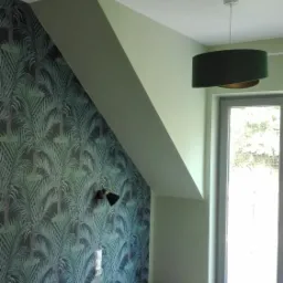Pokój po odświeżeniu: ściana z tapetą w motywy tropikalne, skośny sufit pomalowany na jasnozielono, drewniana podłoga, czarny kinkiet i wisząca lampa z zielonym abażurem.