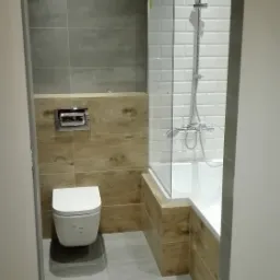 Nowoczesna łazienka po renowacji z szarą podłogą i ścianami, drewnopodobnymi panelami, białą toaletą podwieszaną i wanną z kabiną prysznicową z białymi płytkami.