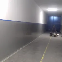 Korytarz w trakcie malowania: dolna część ścian w kolorze szarym, górna biała, odcięcie zabezpieczone niebieską taśmą malarską, widoczne narzędzia malarskie i drabina w tle.