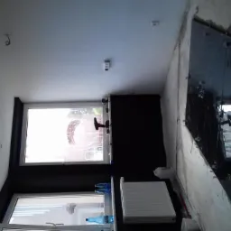 Montaż dużych, ciemnych płytek na ścianie, z użyciem przyssawek i sznurków, widoczne okna i grzejnik w pomieszczeniu w trakcie prac wykończeniowych.