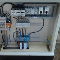 Otwarta szafka elektryczna z zainstalowanymi bezpiecznikami, stycznikami, wyłącznikami i przewodami, widoczne oznaczenia na elementach, instalacja w trakcie montażu.