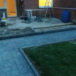 Ułożona kostka brukowa w ogrodzie, widoczna krawędź oddzielająca trawnik, w tle przygotowane materiały i narzędzia do dalszych prac, w tym drabina i beczka.