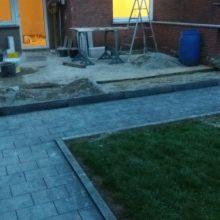 Ułożona kostka brukowa w ogrodzie, widoczna krawędź oddzielająca trawnik, w tle przygotowane materiały i narzędzia do dalszych prac, w tym drabina i beczka.