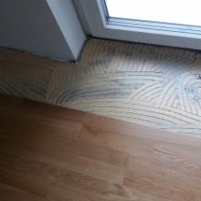 Przygotowanie podłoża pod płytki: warstwa kleju z charakterystycznymi śladami kielni na części powierzchni, sąsiadująca z ułożoną drewnianą podłogą w pobliżu progu okiennego.