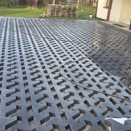 STAN-BRUK - Układanie ażurowej kostki brukowej na podsypce piaskowej. Częściowo ułożona nawierzchnia, widoczny wąż ogrodowy i pracownik w trakcie pracy. Palety w tle.