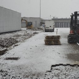 STAN-BRUK - Zimowy krajobraz placu budowy z koparką w tle, paletami na pierwszym planie i padającym śniegiem. Widoczny fragment utwardzonego terenu i biały bus.