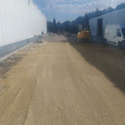 STAN-BRUK - Żółty minikoparka CAT i zagęszczarka na placu budowy, przygotowany teren pod budowę, widoczne budynki magazynowe i biały van. Praca w toku, słoneczny dzień.