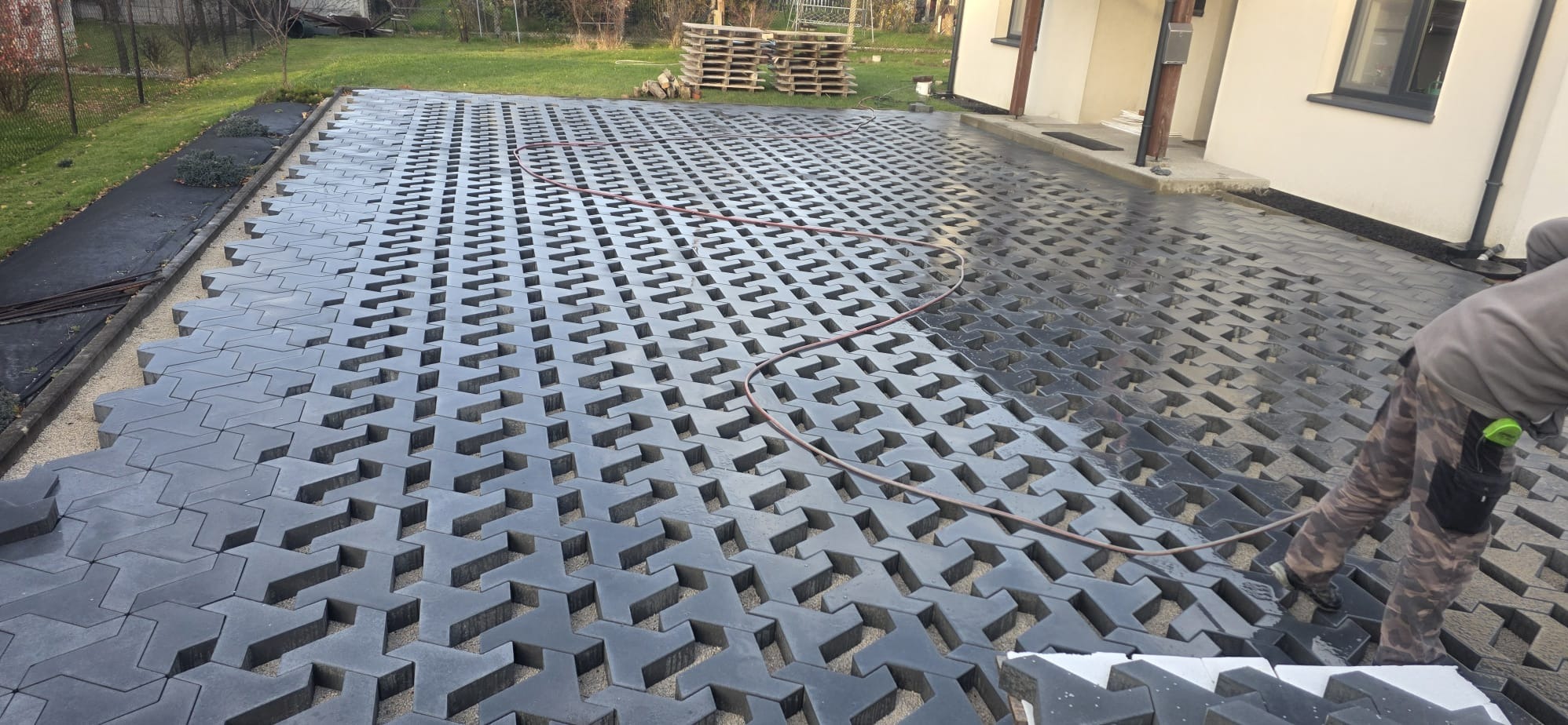 Układanie ażurowej kostki brukowej na podsypce piaskowej. Częściowo ułożona nawierzchnia, widoczny wąż ogrodowy i pracownik w trakcie pracy. Palety w tle.