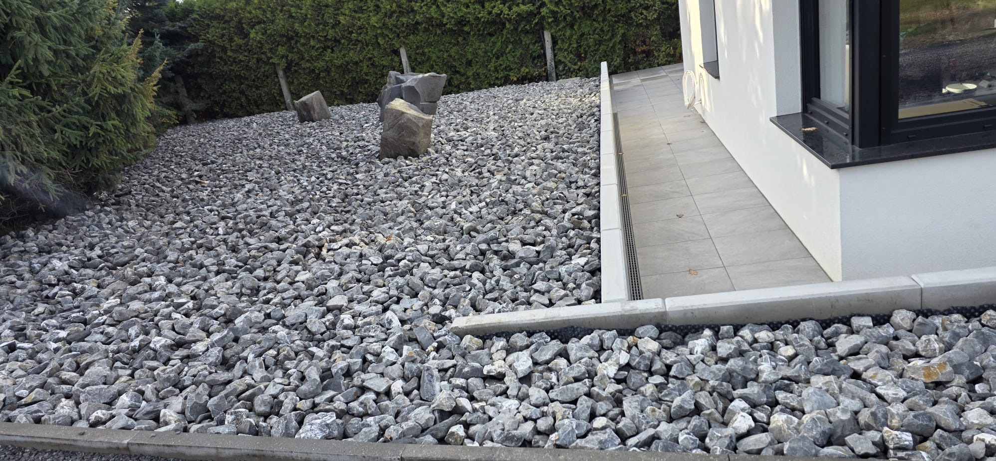Szare kamienie dekoracyjne w ogrodzie przy nowoczesnym domu z jasną elewacją i ciemnymi oknami. Ułożone kamienie tworzą estetyczny wygląd.