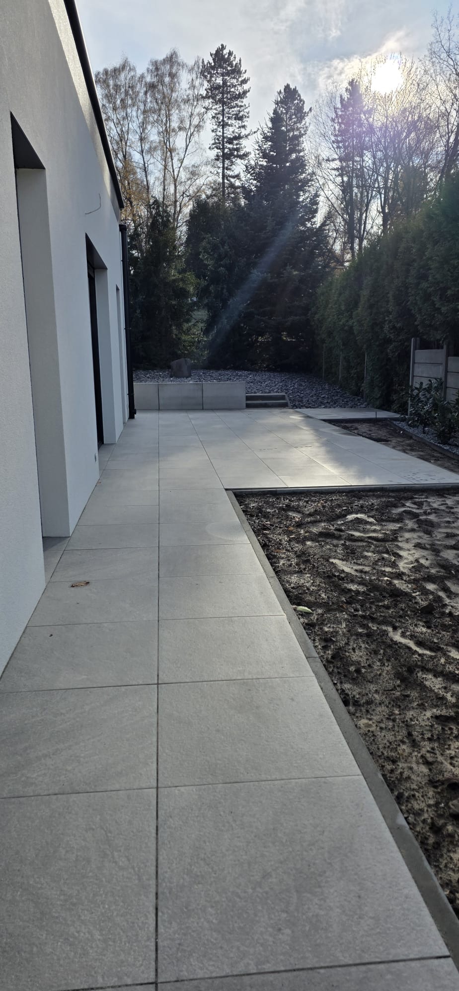 Nowoczesny taras z jasnoszarej kostki brukowej, przylegający do białego budynku. Widoczne drzewa w tle i przygotowany teren pod nasadzenia.