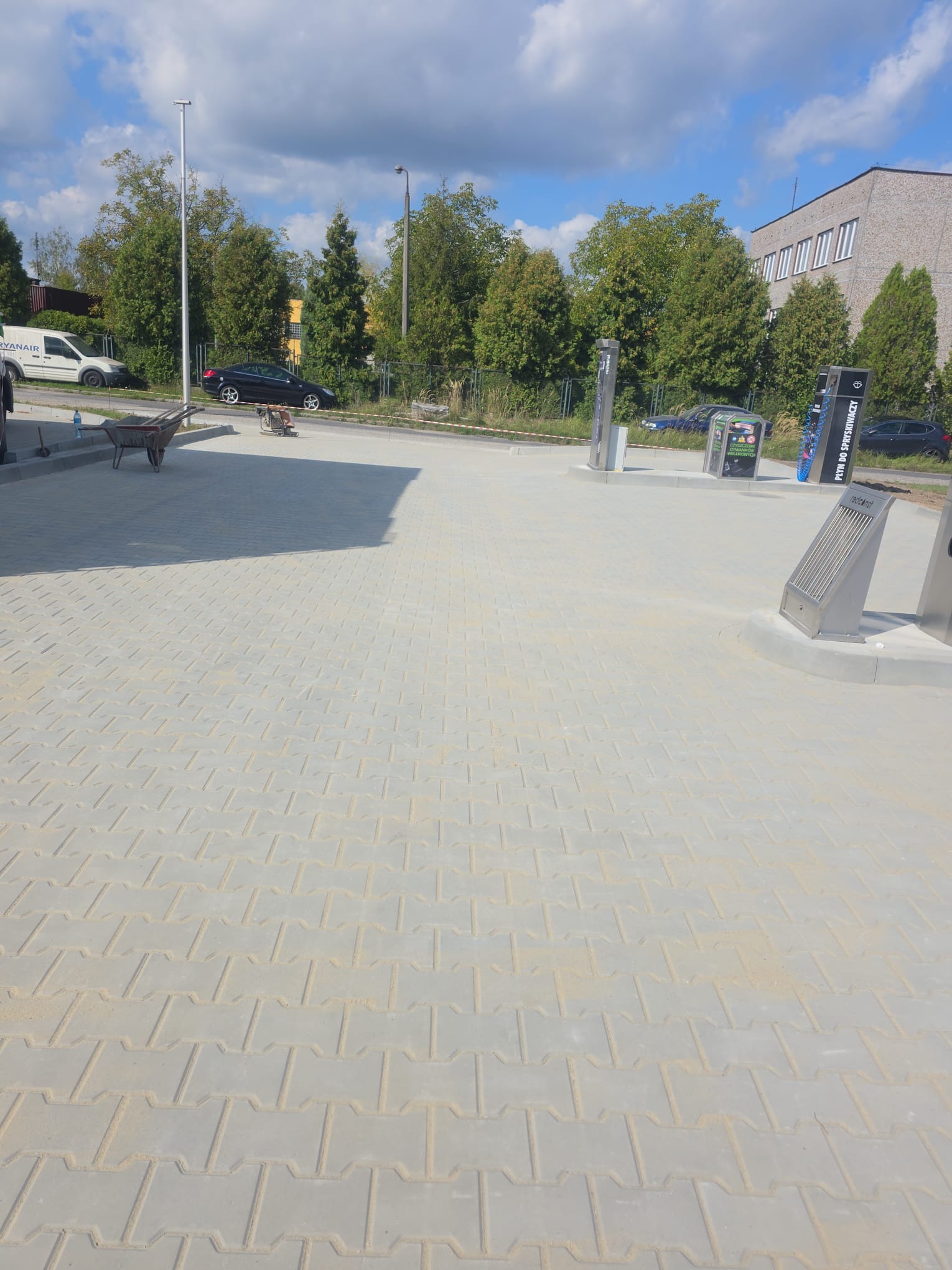 Jasnoszara kostka brukowa ułożona na dużej powierzchni, widoczny wózek i maszyna do zagęszczania, drzewa w tle. Nowa nawierzchnia z kostki brukowej.