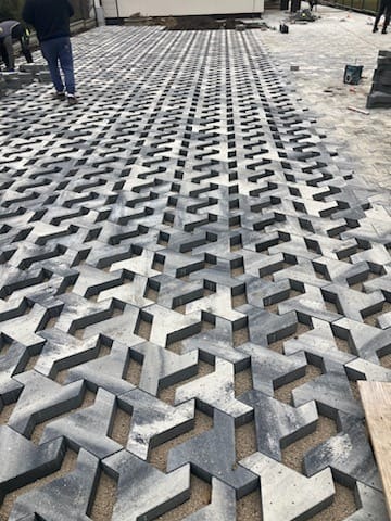 Szara, ażurowa kostka brukowa ułożona w geometryczny wzór na podjeździe. Widoczne piaskowe wypełnienie i fragmenty niezakończonej powierzchni. Perspektywa z dołu do góry.