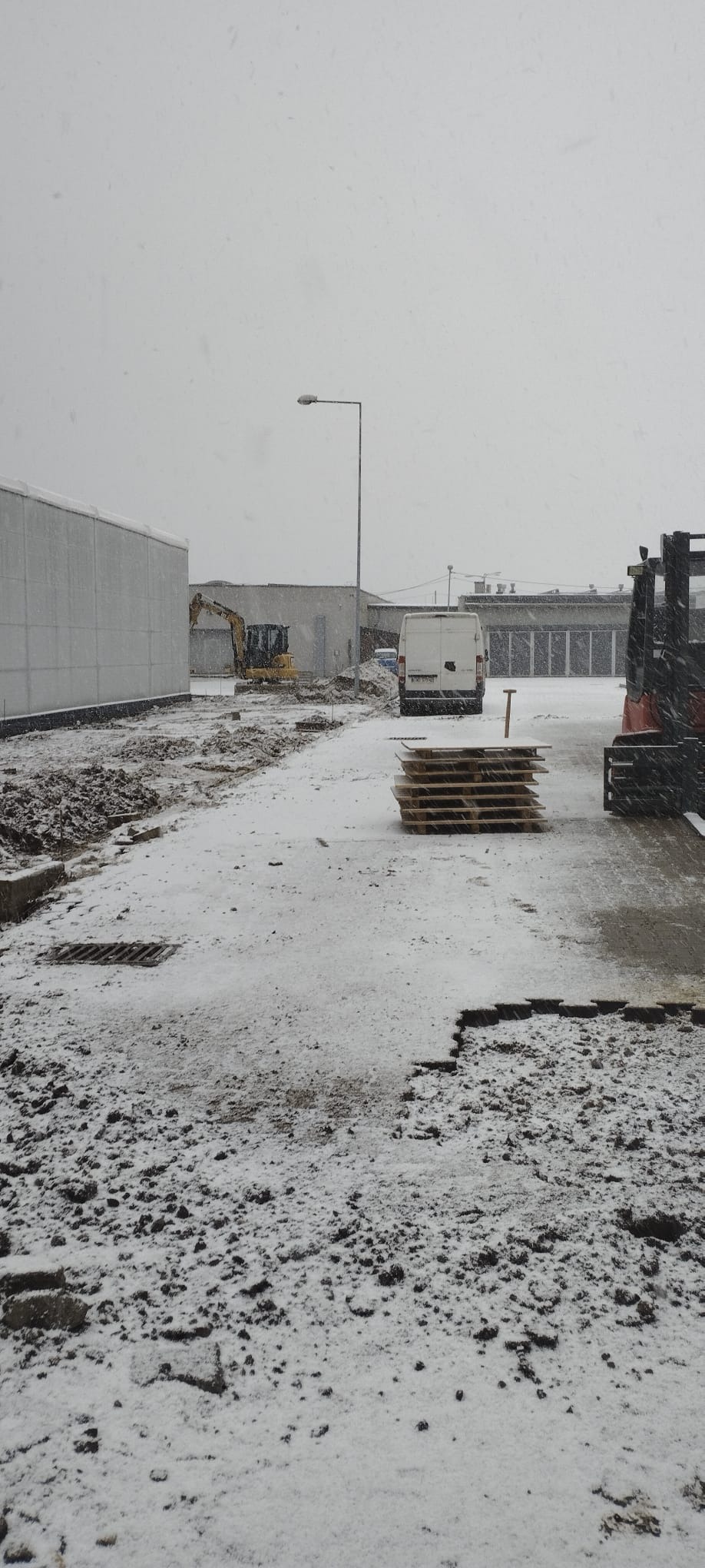 Zimowy krajobraz placu budowy z koparką w tle, paletami na pierwszym planie i padającym śniegiem. Widoczny fragment utwardzonego terenu i biały bus.