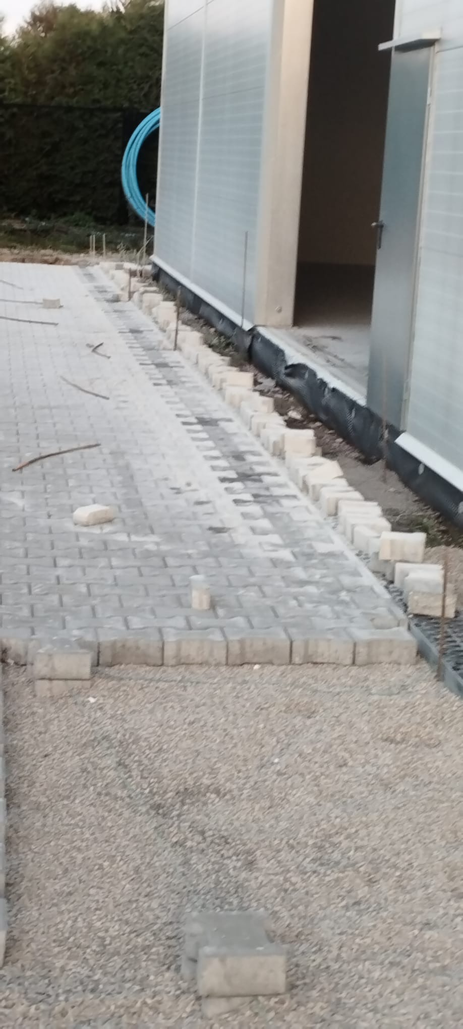 Układanie szarej kostki brukowej na podsypce, widoczne niedokończone obrzeża i fragment budynku z blachy trapezowej w tle. Prace brukarskie w toku.