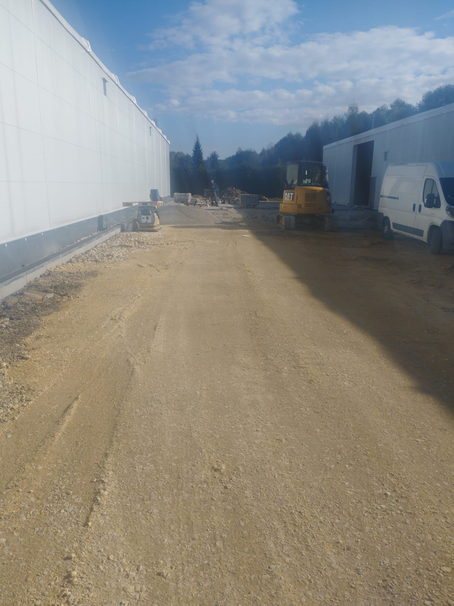 Żółty minikoparka CAT i zagęszczarka na placu budowy, przygotowany teren pod budowę, widoczne budynki magazynowe i biały van. Praca w toku, słoneczny dzień.