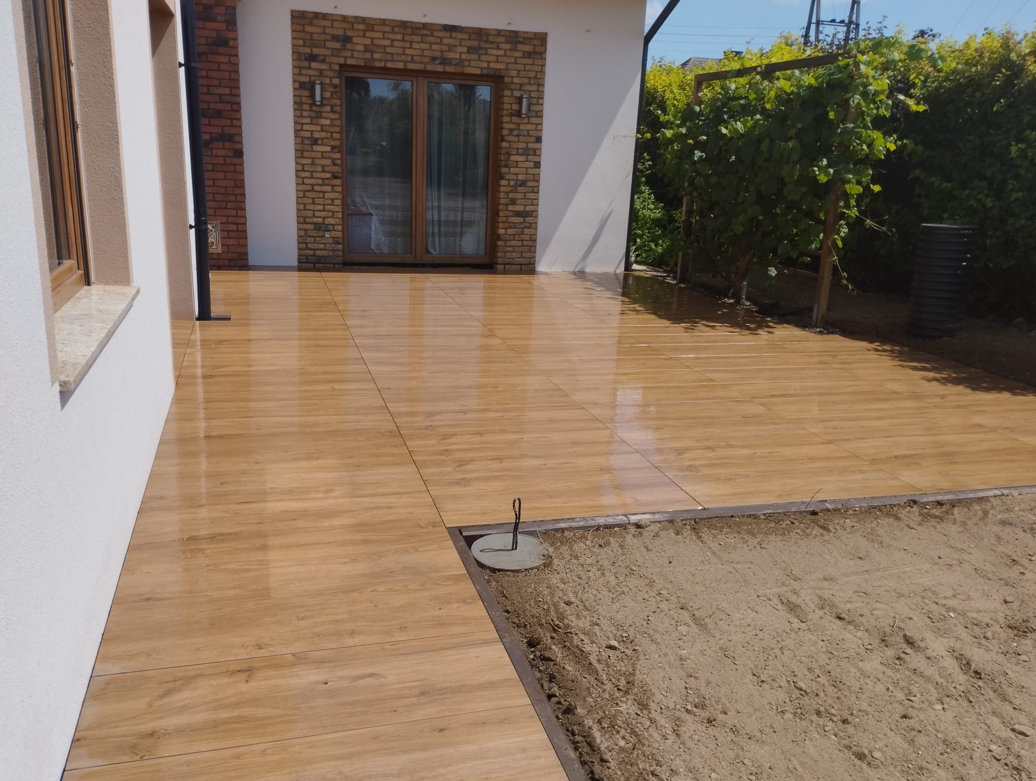 Błyszczący taras z płytek imitujących drewno, z widocznym fragmentem przygotowanego podłoża pod dalsze prace. Ujęcie z niskiej perspektywy podkreśla detale wykończenia.