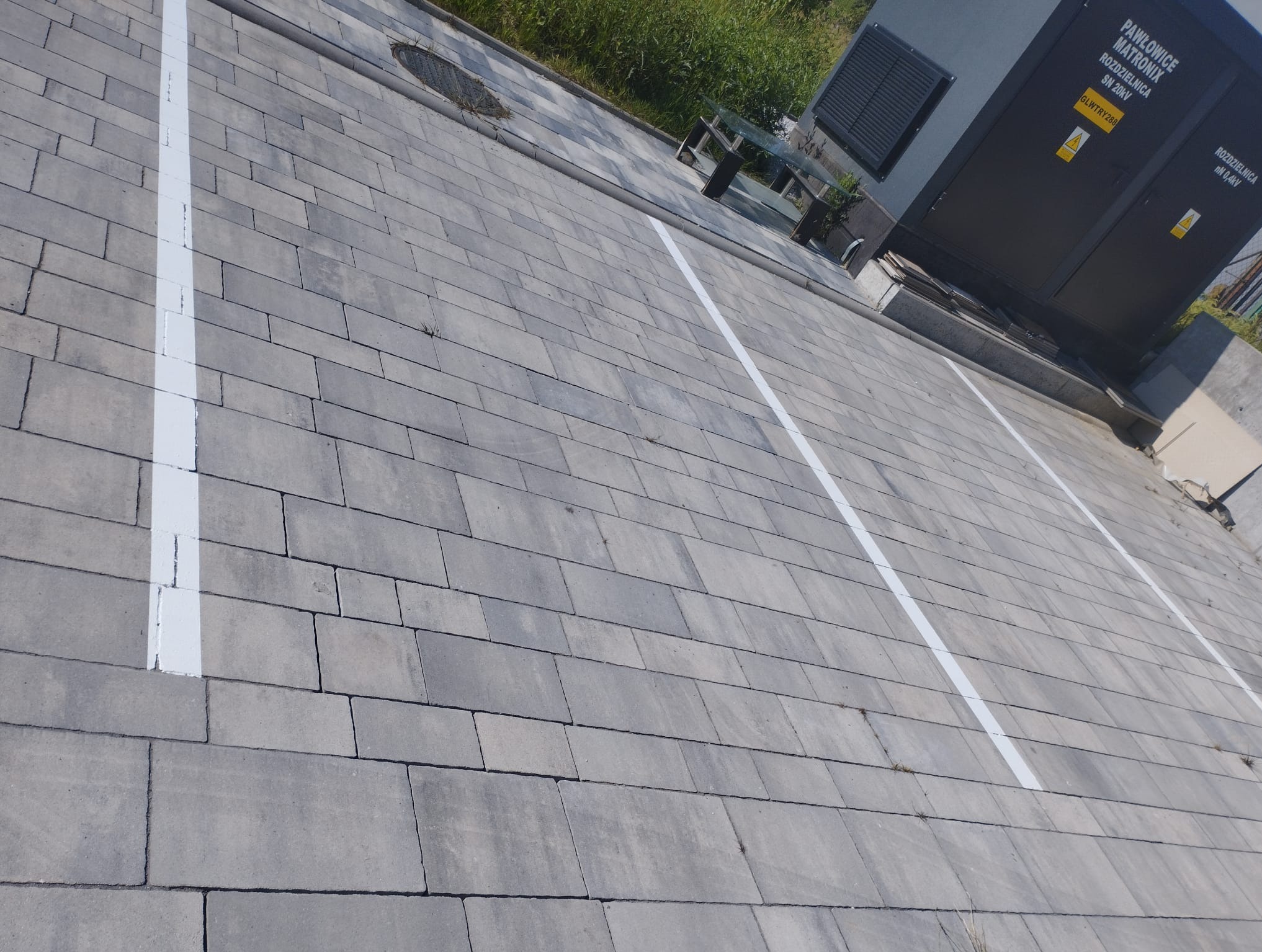 Szara kostka brukowa z białymi liniami wyznaczającymi miejsca parkingowe, widok z góry, obok transformator i trawa.