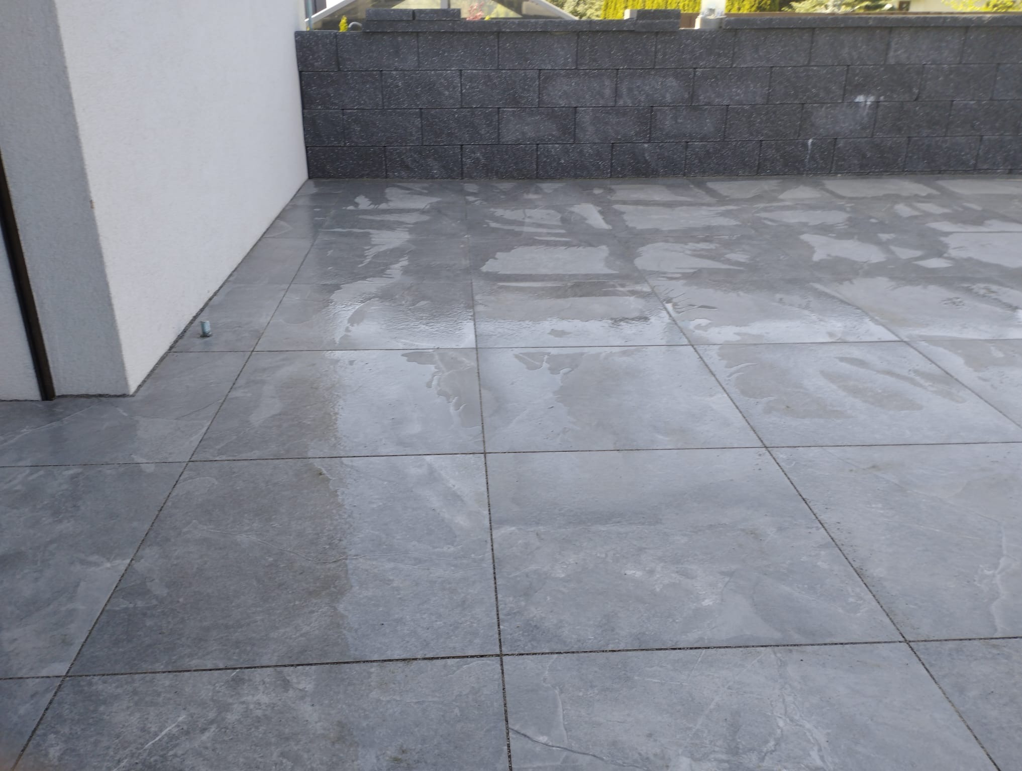 Szara, kwadratowa kostka brukowa ułożona na zewnątrz, przylegająca do białej ściany i niskiego murku z ciemnoszarej kostki. Widoczne delikatne refleksy wody na powierzchni.