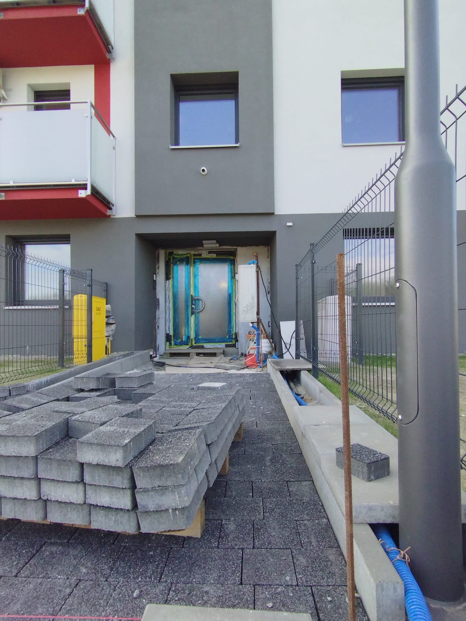 Układanie szarej kostki brukowej przed nowoczesnym budynkiem. Częściowo ułożona nawierzchnia, paleta z kostką, widoczne elementy drenażu i ogrodzenie.