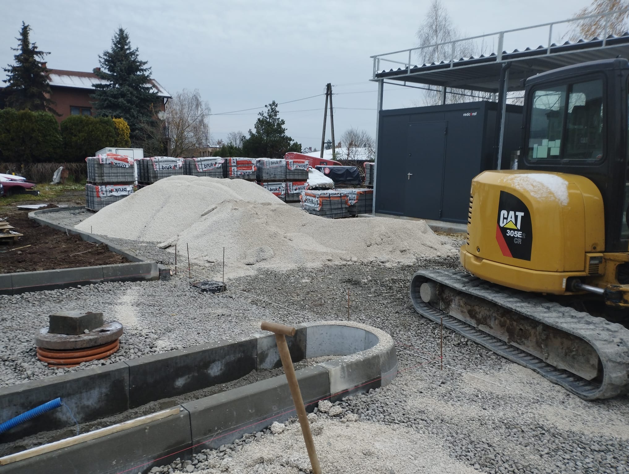Przygotowanie terenu pod kostkę brukową: krawężniki, podsypka, mini koparka CAT 305E CR i palety kostki brukowej. Widok na plac budowy w pochmurny dzień.