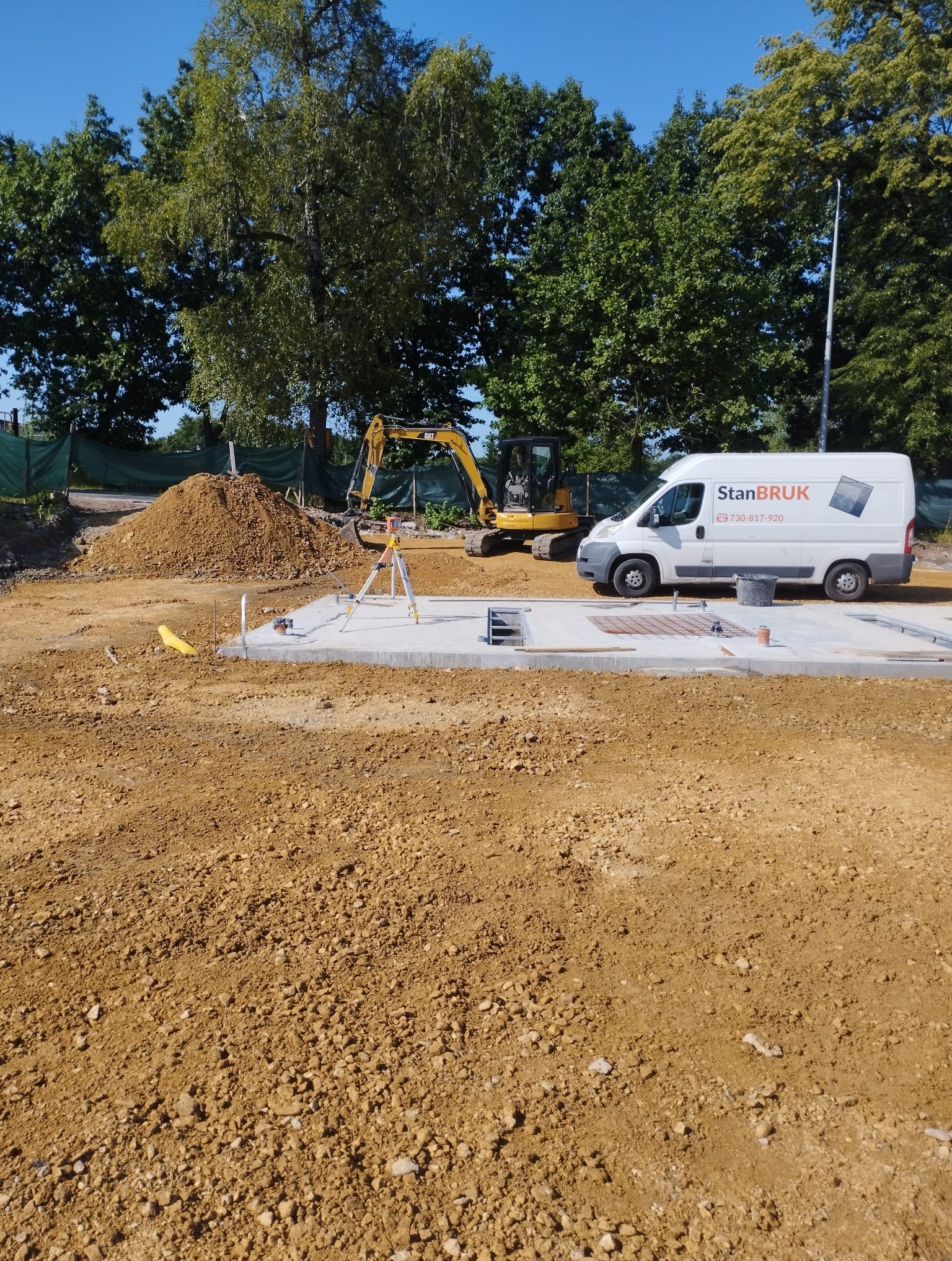 Przygotowany teren pod fundamenty z widocznym zbrojeniem, niwelatorem, koparką i samochodem dostawczym firmy StanBRUK w słoneczny dzień.