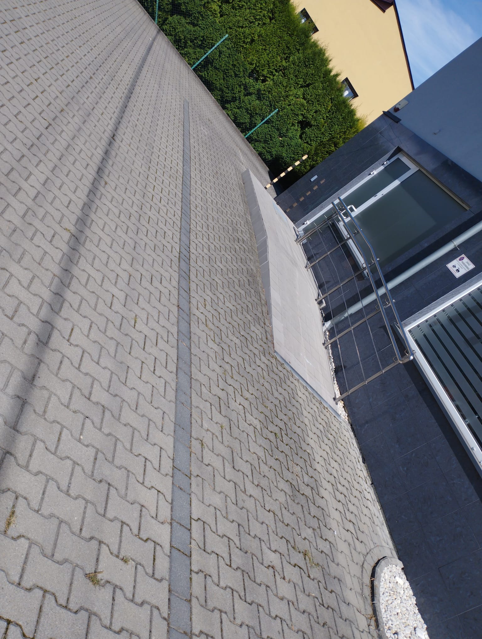 Szara kostka brukowa ułożona wzorem jodełki, oddzielona pasem z ciemniejszych kostek. Widok z góry, obok budynku z szarymi płytami i metalowymi schodami.