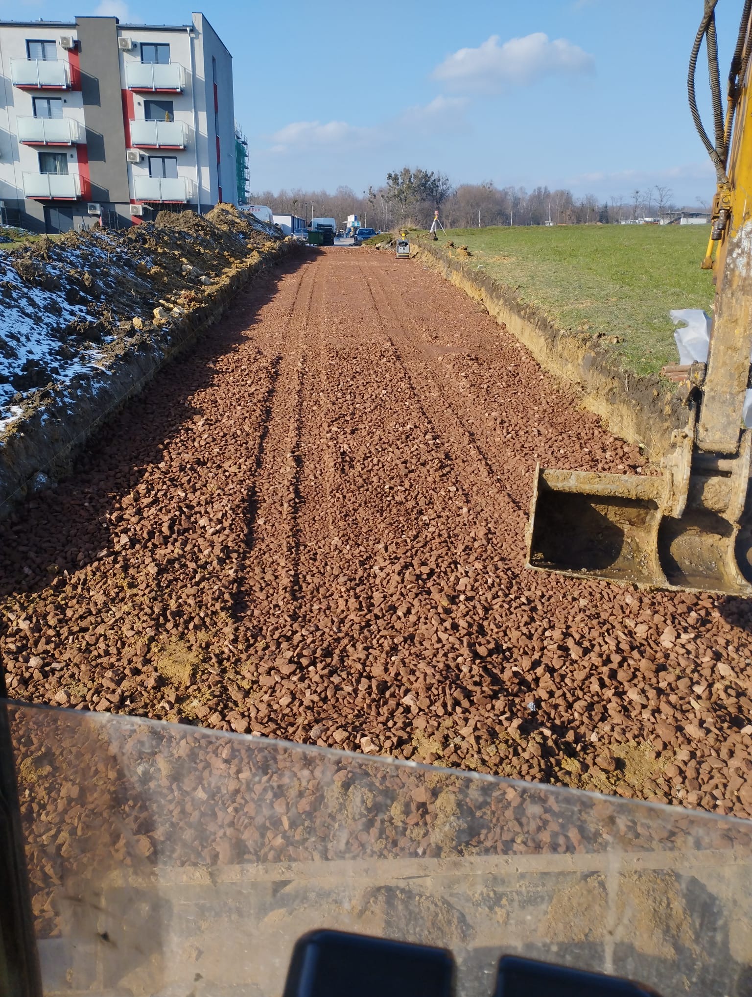 Wykop pod fundamenty budynku wielorodzinnego, wypełniony czerwonym gruzem. Widoczny fragment koparki i budynek w tle. Prace ziemne w toku, teren przygotowany pod budowę.