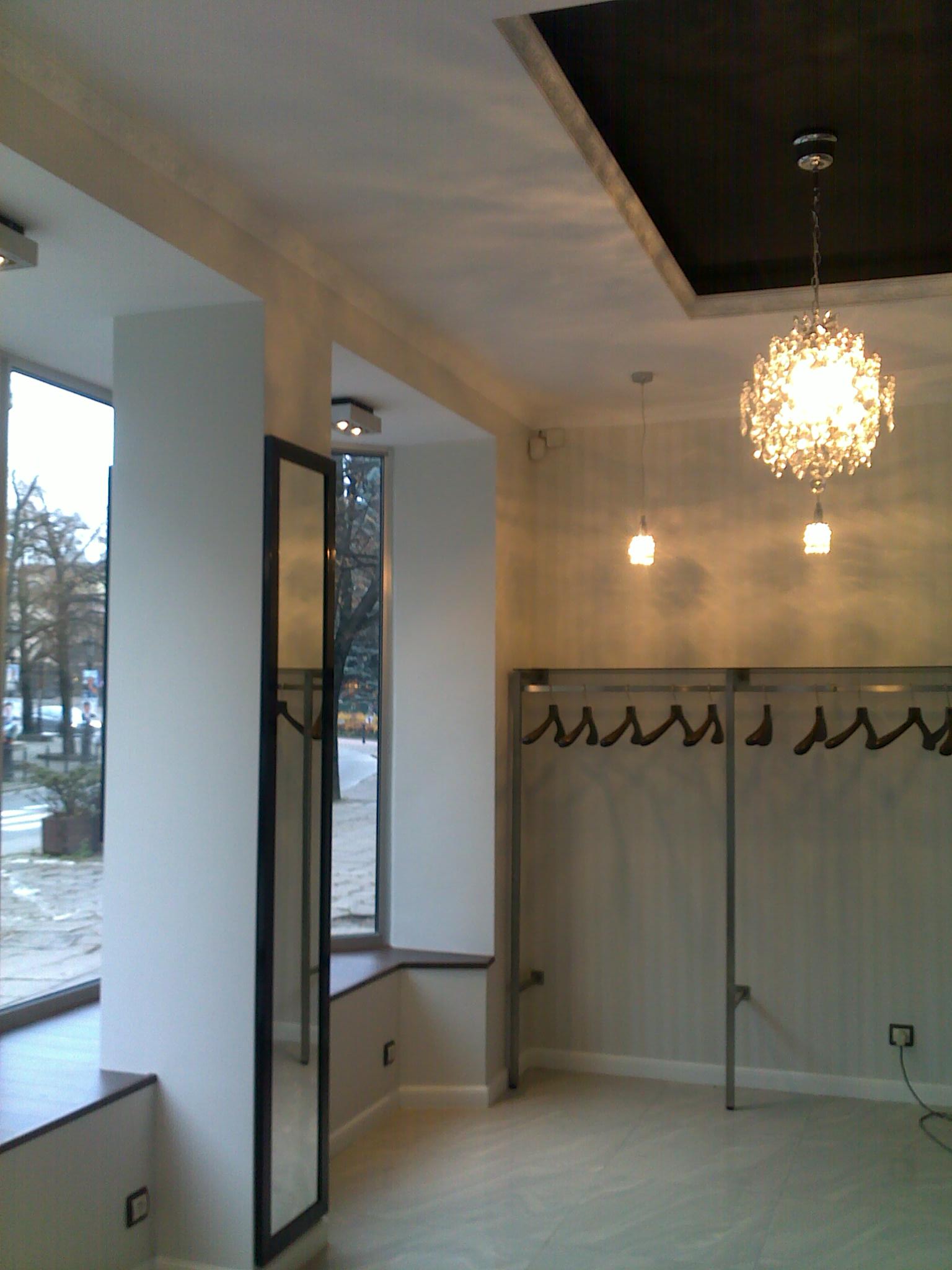 Pusta garderoba w jasnym pomieszczeniu z dużymi oknami, wiszącymi wieszakami i ozdobnym żyrandolem.