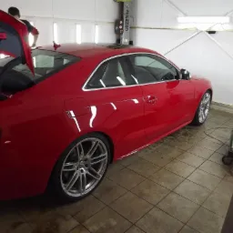 Czerwone auto sportowe z otwartym bagażnikiem na podnośniku w warsztacie, widoczne aluminiowe felgi i odbicie światła w lakierze.