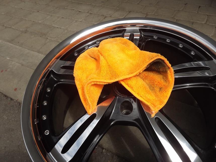 Błyszcząca, czarno-srebrna felga aluminiowa po czyszczeniu, z pomarańczową ściereczką z mikrofibry na wierzchu, leżąca na szarym chodniku.