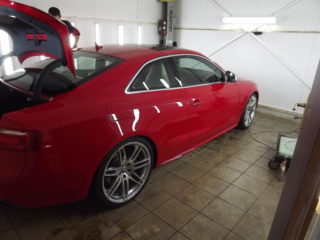 Czerwone auto sportowe z otwartym bagażnikiem na podnośniku w warsztacie, widoczne aluminiowe felgi i odbicie światła w lakierze.