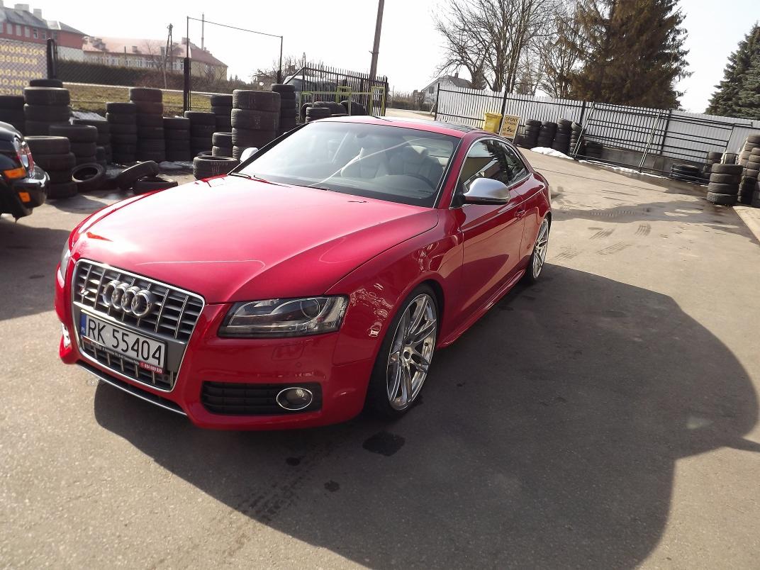 Czerwone Audi S5 z chromowanymi felgami, zaparkowane na placu z oponami w tle, widoczne tablice rejestracyjne.