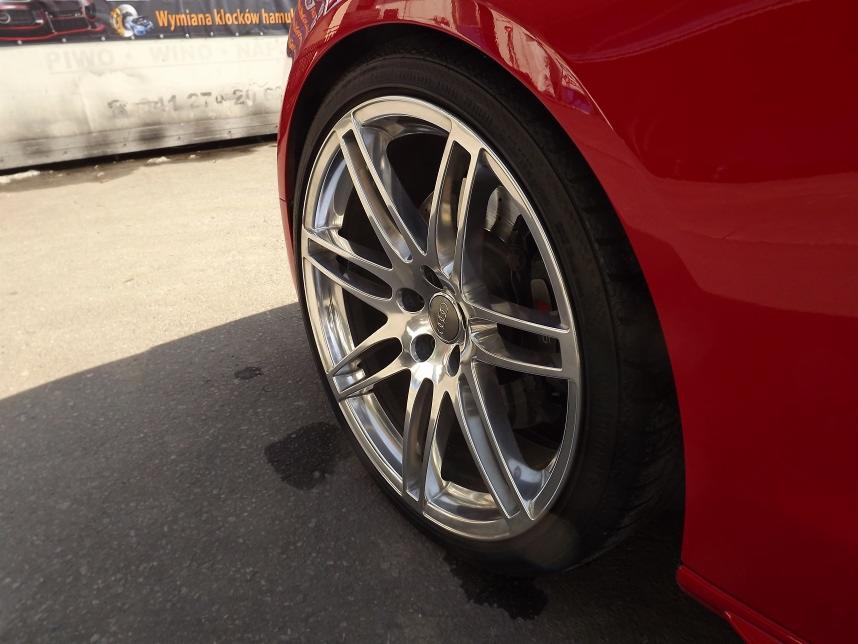 Błyszcząca, aluminiowa felga z oponą na czerwonym samochodzie marki Audi, widok z boku, na tle reklamy warsztatu.