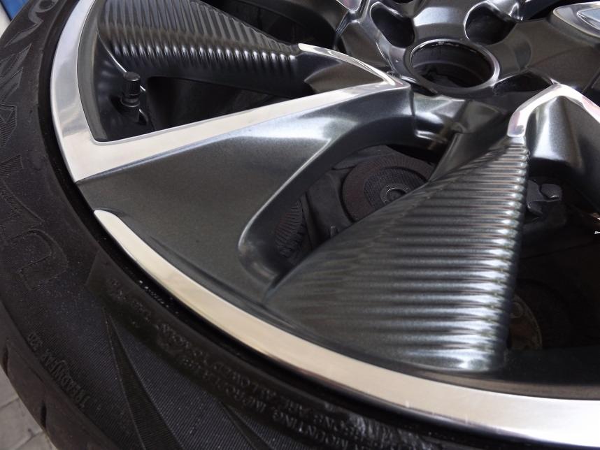 Błyszcząca, ciemnoszara felga aluminiowa z oponą, widoczne detale bieżnika i zacisk hamulcowy w tle.