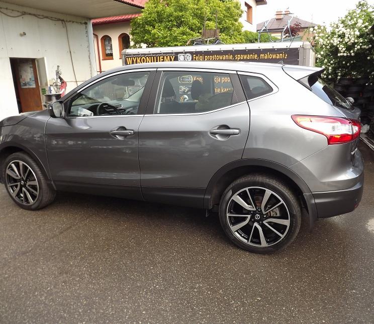 Szary Nissan Qashqai po renowacji felg, widoczny na zewnątrz warsztatu z reklamą usług prostowania, spawania i malowania felg.
