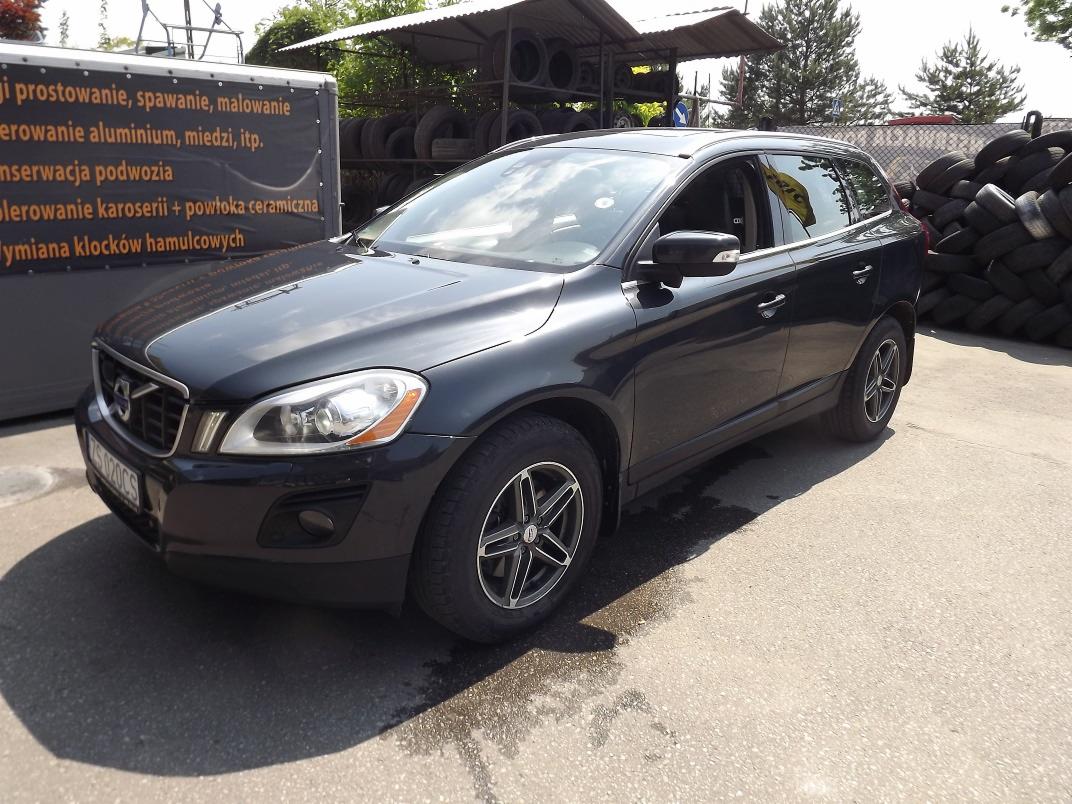 Czarny samochód Volvo XC60 zaparkowany na betonowym placu, z widoczną przyczepą reklamową w tle i stertą opon, prawdopodobnie po serwisie.