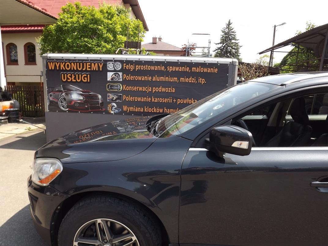 Ciemny samochód osobowy zaparkowany przed przyczepą reklamującą usługi warsztatowe: prostowanie felg, polerowanie aluminium, konserwacja podwozia, polerowanie karoserii i wymiana klocków hamulcowych.