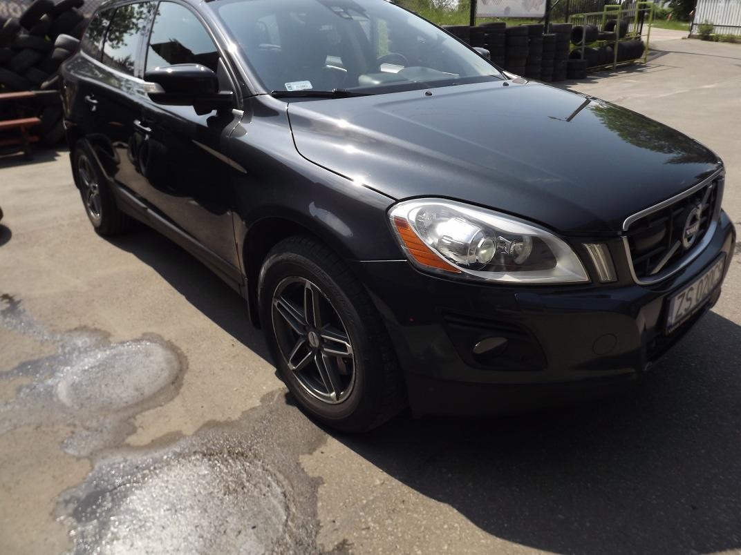 Czarny samochód Volvo XC60 po umyciu, z widocznymi śladami wody na karoserii i mokrym podłożu, zaparkowany na placu w pobliżu sterty opon.