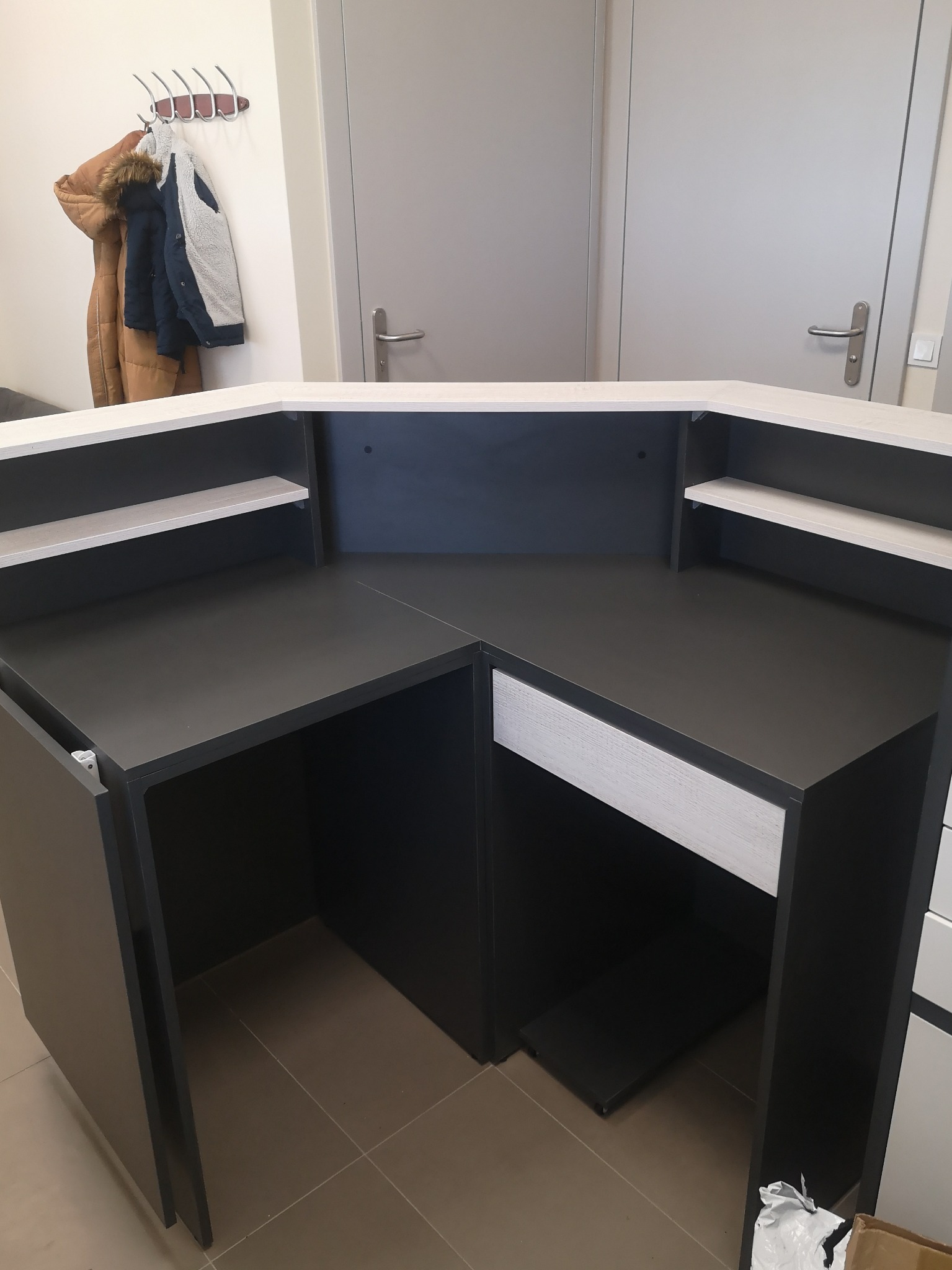 Nowoczesna lada recepcyjna w kolorze grafitowym z elementami w odcieniu jasnego drewna, widoczne półki i szuflada, na ścianie wieszak z kurtkami.