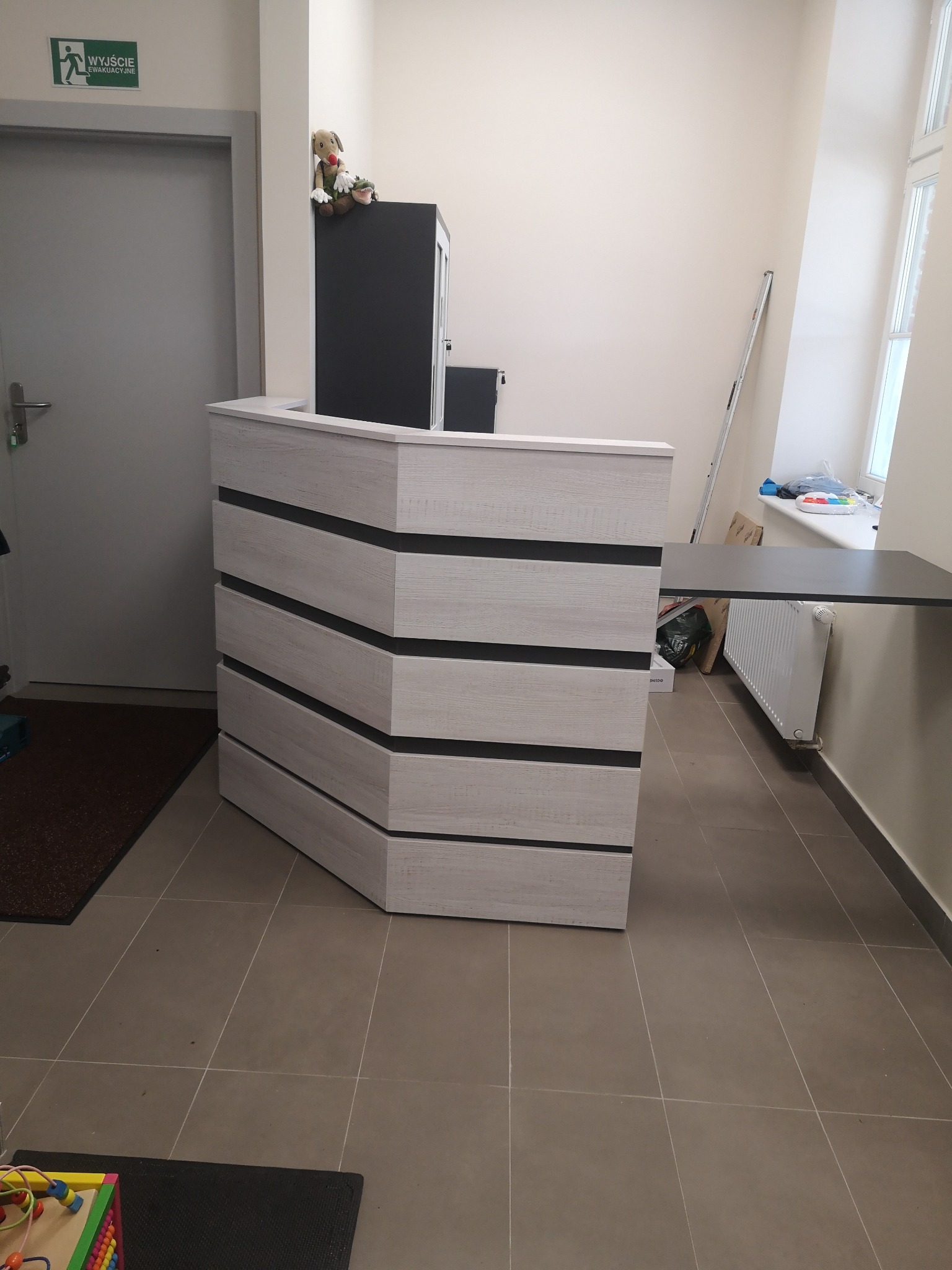 Nowoczesna lada recepcyjna z drewnopodobną fakturą, z czarnymi poziomymi pasami, ustawiona w jasnym pomieszczeniu z oknem i grzejnikiem, z zabawkami dla dzieci w pobliżu.