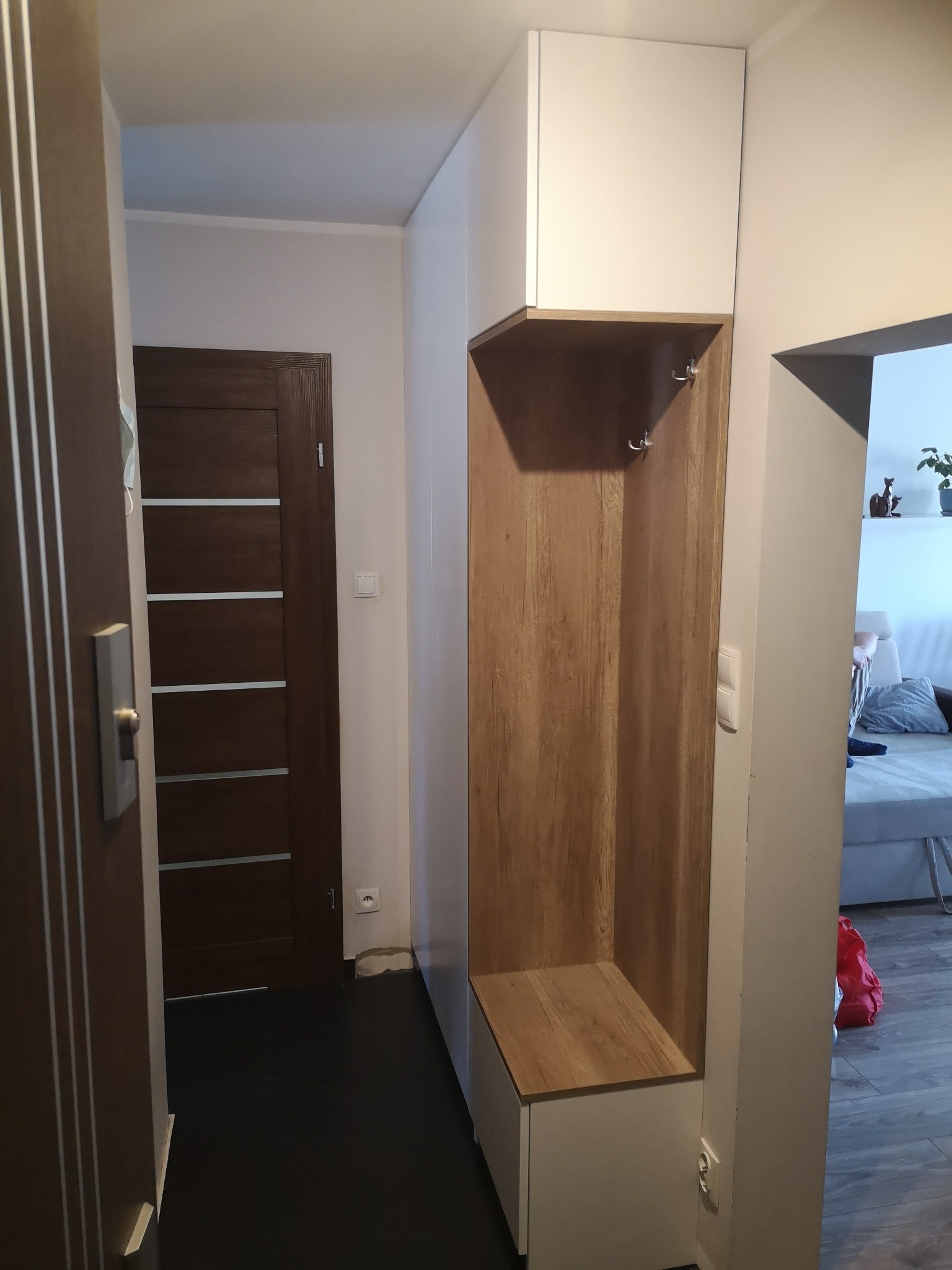 Wnęka z półką i wieszakami w przedpokoju, wykończona białymi płytami i okleiną imitującą drewno, widok na drzwi i fragment salonu.