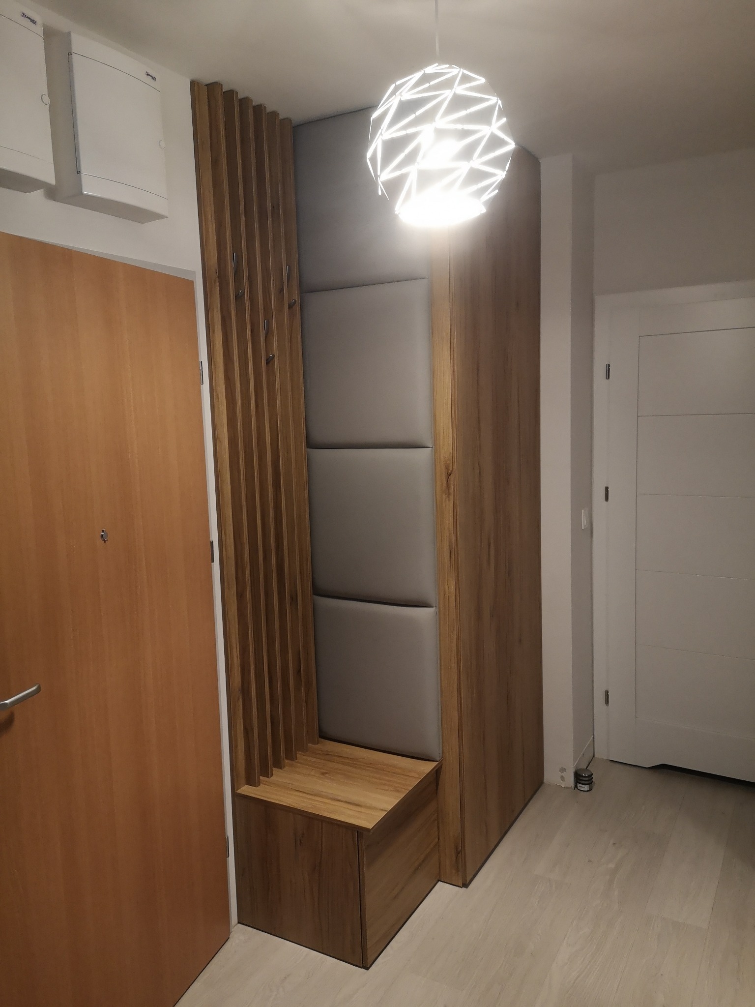 Nowoczesna szafa wnękowa z siedziskiem i panelem tapicerowanym, oświetlona dekoracyjną lampą wiszącą w przedpokoju, z drewnianymi elementami i wieszakami.