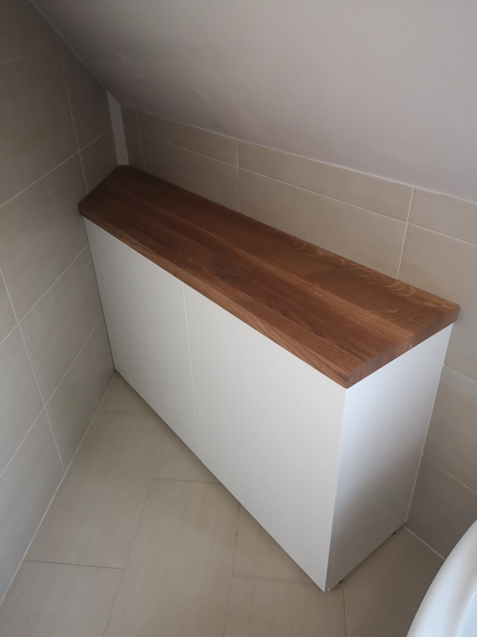 Biała szafka łazienkowa pod skosem dachu, z blatem z jasnego drewna, dopasowana do beżowych płytek na ścianie i podłodze.