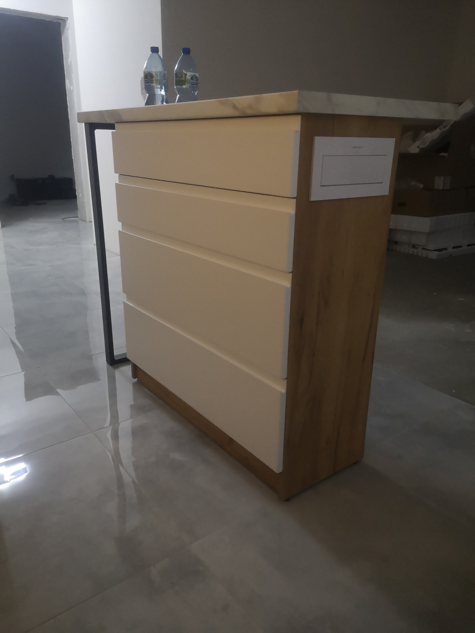 Niska komoda z czterema szufladami w kolorze kremowym, z blatem imitującym marmur, stoi na błyszczącej, szarej podłodze. Widoczne dwie butelki wody na blacie i surowy stan wykończenia wnętrza.