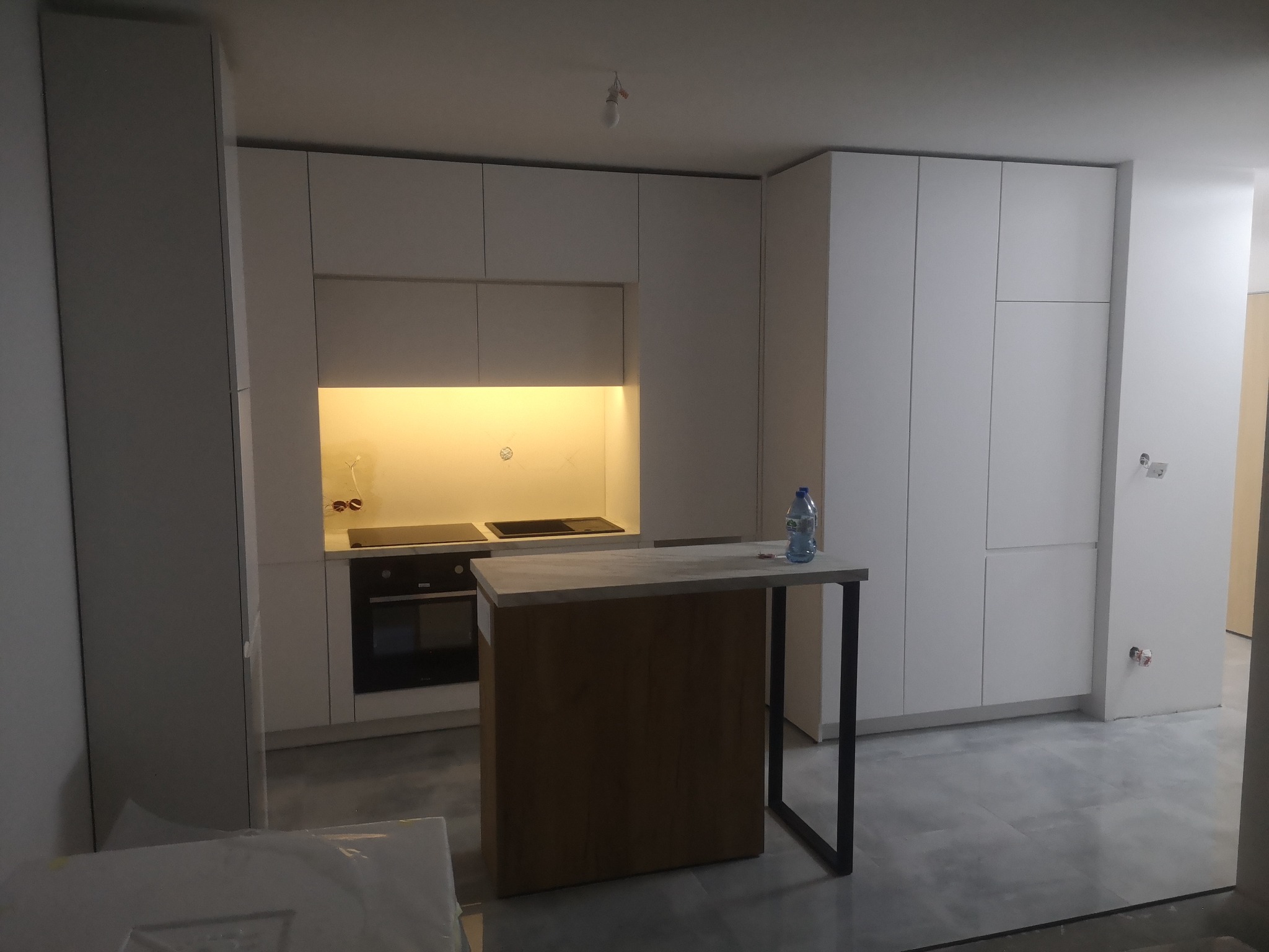 Nowoczesna, minimalistyczna kuchnia z białymi frontami szafek, oświetleniem podszafkowym i wyspą z blatem betonowym oraz czarnymi nogami. Widoczna płyta indukcyjna i piekarnik. Na blacie wyspy stoi...