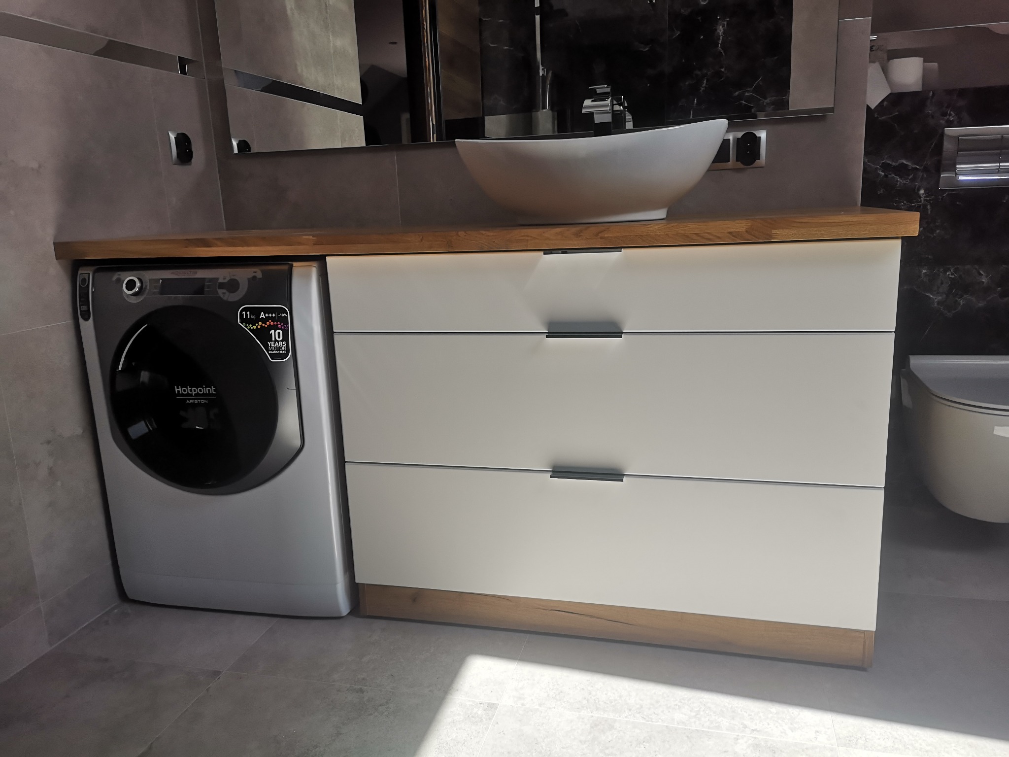 Nowoczesna łazienka z pralką Hotpoint Ariston w zabudowie szafki pod umywalką nablatową, trzy szuflady z minimalistycznymi uchwytami, szara marmurowa ściana w tle.