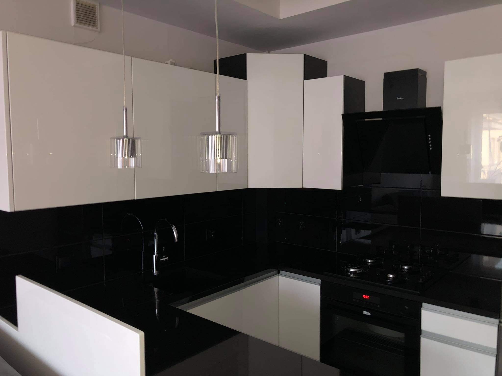 Nowoczesna aranżacja kuchni w Czerwonaku z białymi, lakierowanymi frontami szafek, czarnym blatem i błyszczącymi płytkami na ścianie, oświetlona wiszącymi lampami z chromowanymi detalami.