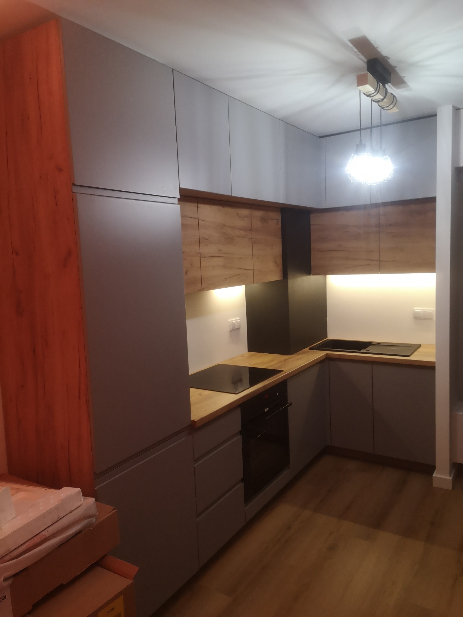 Nowoczesna kuchnia z szarymi frontami szafek i drewnianymi akcentami, oświetlona listwami LED pod szafkami wiszącymi i designerską lampą sufitową; w tle częściowo widoczne kartony z materiałami...